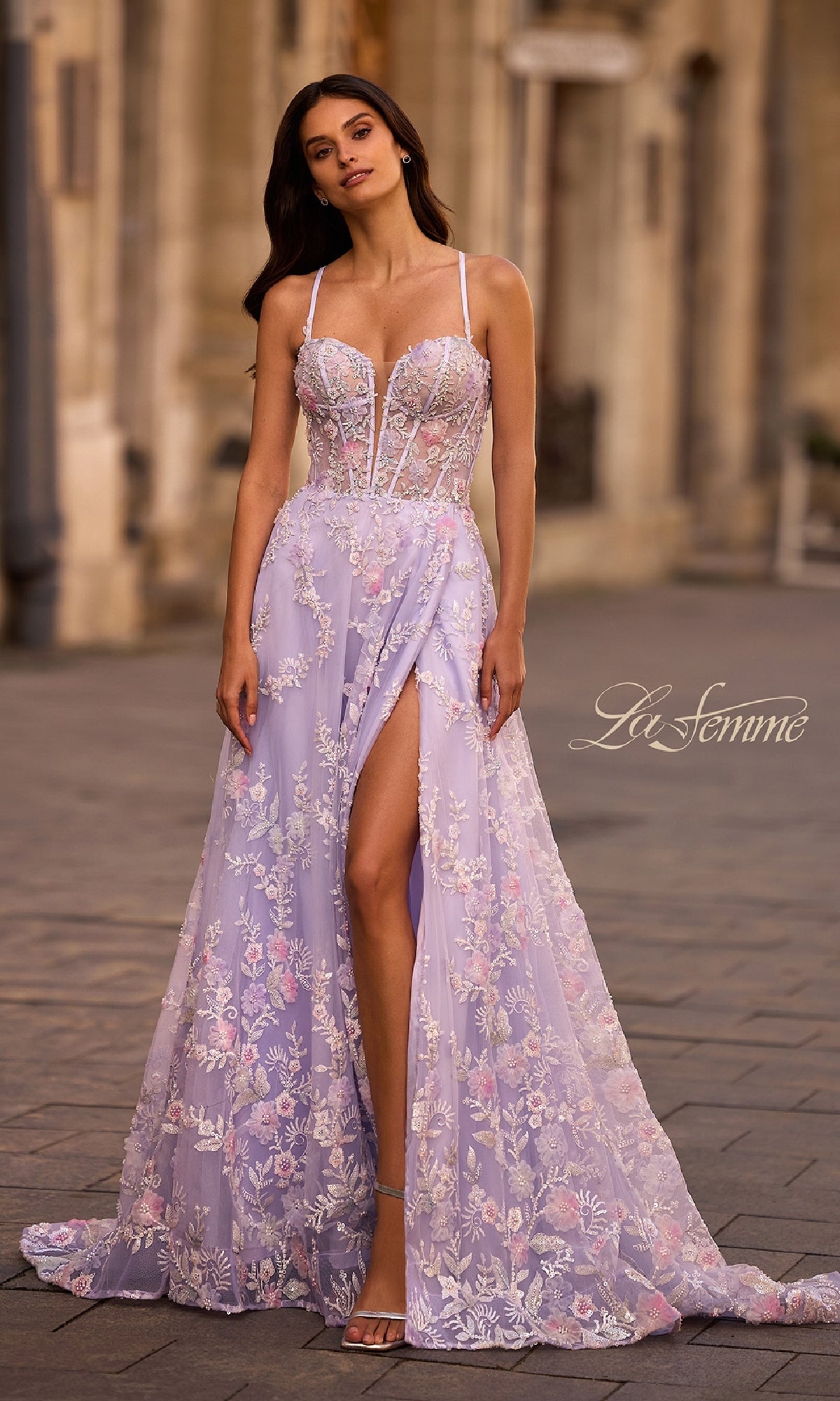 A woman stands on a city street in the La Femme 32695 long prom dress—a lavender gown featuring floral lace embroidery, a sheer bodice, thin straps, and a high slit.